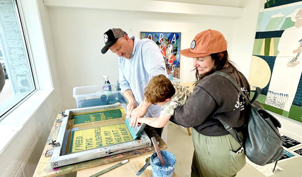 Adam Bridgland helping a young boy to screenprint. The boy is being held up by his smiling mum.